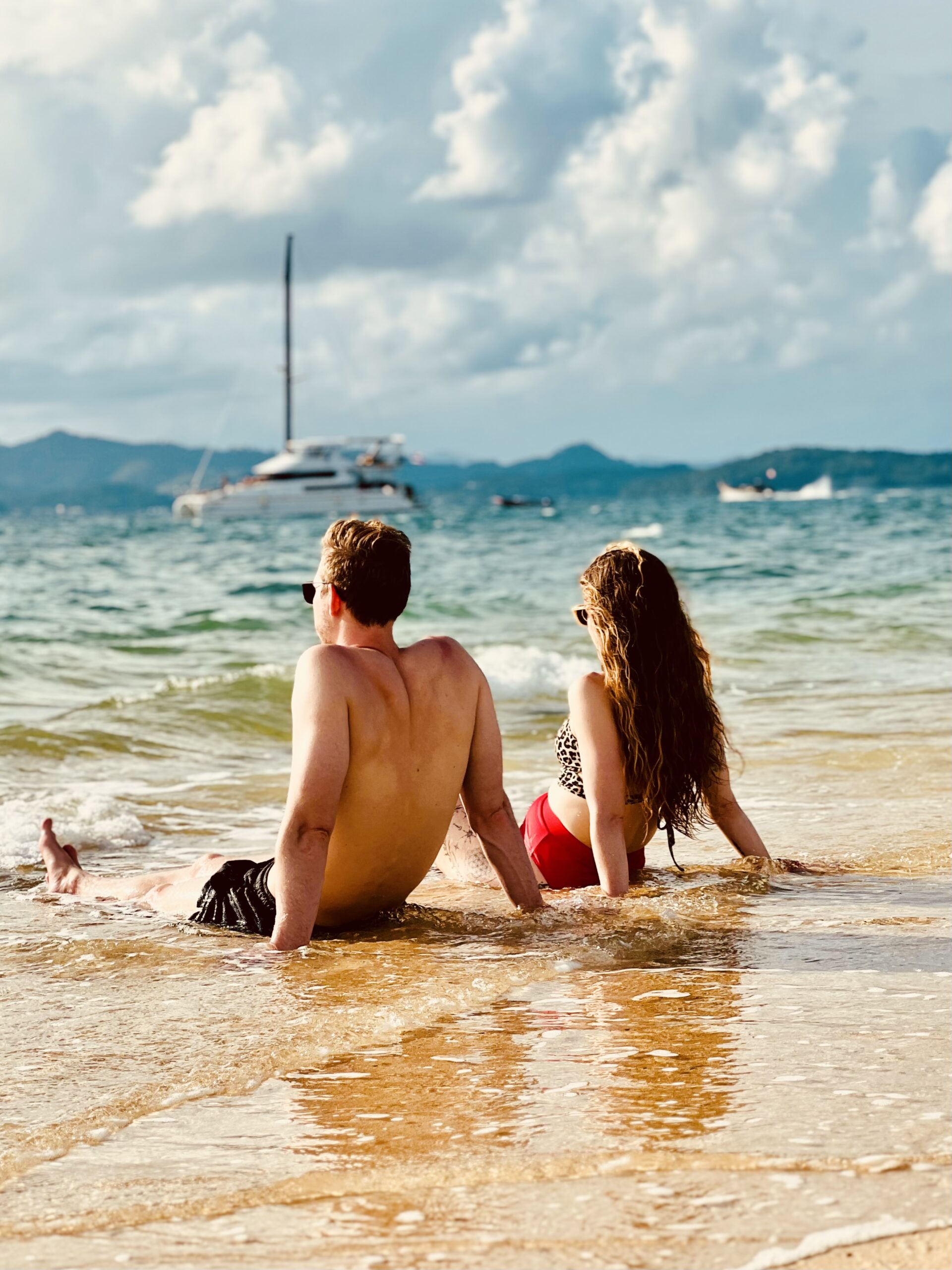 Thailand Island Boat Day Couple sitting in shallow water looking out at a catamaran near the islands in Thailand